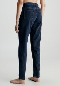 Barfota person som bär mörkblå högmidjade jeans med bakfickor, står mot en ljus, enfärgad bakgrund, sett bakifrån.