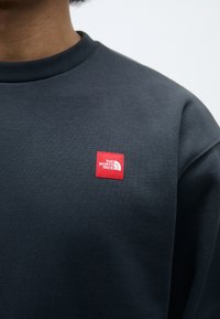 Sudadera gris oscuro con cuello de canalé que presenta un pequeño logo cuadrado rojo con texto blanco y diseño en arco en el lado izquierdo del pecho. Material suave.