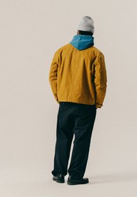 Άτομο που φοράει γκρι σκουφί, μπλε φούτερ, μουσταρδί μπουφάν, μαύρο παντελόνι και μαύρα παπούτσια, στέκεται με την πλάτη γυρισμένη σε μια απλή φόντο.