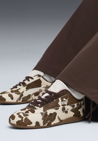 Sapatilhas brancas e castanhas com estampa de vaca, em camurça e pele texturizadas. Cadarços castanhos e logótipo da Puma; usadas com calças castanhas escuras.