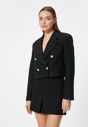 Zwarte cropped blazer van een gestructureerde stof, dubbelzijdig ontwerp en accenten van zilveren knopen. Gecombineerd met een bijpassende mini-rok met een split aan de voorkant.