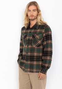 Mand med langt blond hår iført en grøn, sort og beige ternet skjorte med knapper og beige bukser, stående med den ene hånd i lommen.