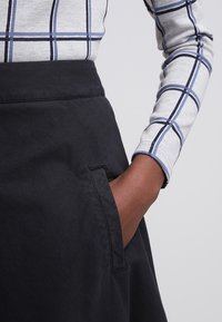 Jupe noire avec une large ceinture et une poche latérale, associée à une chemise à manches longues grise avec un motif en grille bleue. Texture de tissu lisse.