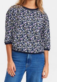 Frau in einer marineblauen Bluse mit Blumenmuster und 3/4-Ärmeln, trägt blaue Jeans und zeigt Oberkörper und Oberschenkel, mit einem goldenen Armband am linken Handgelenk.