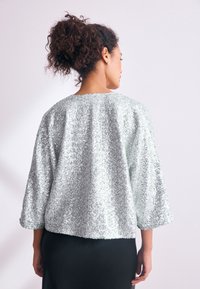 Silberne Paillettenjacke mit Dreiviertelärmeln, rundem Ausschnitt und lockerem Schnitt, von hinten vor hellem Hintergrund gezeigt.