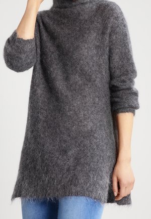 Personne portant un pull à col roulé long, duveteux et gris avec les manches retroussées, associé à un jean bleu, sur un fond clair uni.
