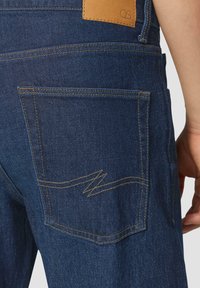 Salopette en denim bleu foncé avec une étiquette en cuir et un détail de couture en zigzag sur la poche arrière. Couture visible dans une couleur contrastante.