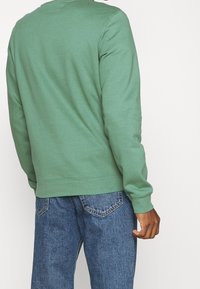 Sudadera verde de tejido suave, con cuello redondo, mangas largas y puños de canalé. Combinada con vaqueros azules.