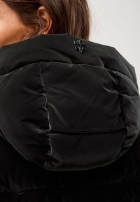 Zwarte pufferjack met een doorgestikt ontwerp en glanzende afwerking, met een verhoogd logo-detail op de capuchon aan de achterkant.