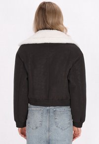 Schwarze Cropped-Jacke mit einem weißen Kunstpelzkragen, strukturiertem Stoff, elastischem Bund und Bündchen, kombiniert mit hellblauen Jeans.
