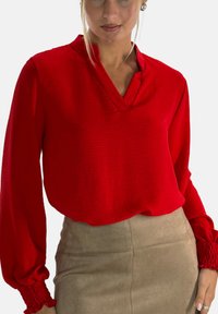 Red long-sleeve blouse with a V-neckline and textured fabric, featuring gathered cuffs and a relaxed fit, paired with a beige skirt.