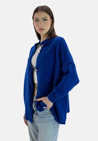 Cárdigan azul de punto con cuello redondo, botones delanteros grandes y mangas con puños. Presenta un corte holgado y una textura de tela.