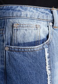 Světle modré džíny z denimu s bledým designem, vybavené přední kapsou, viditelným šitím a ošuntělými okraji u pasu.