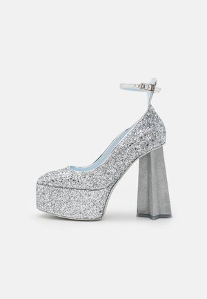 Zapato de tacón alto con plataforma y purpurina plateada, con correa en el tobillo y tacón ancho en bloque cónico sobre fondo blanco.