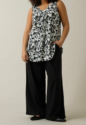 Femme portant un haut sans manches noir et blanc à motifs floraux, un pantalon large noir et des chaussures noires pointues, debout avec une main dans la poche.