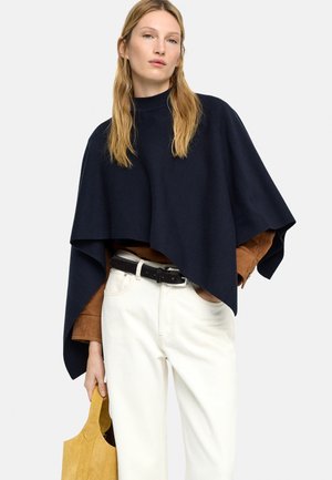 Kobieta w granatowym ponczo na brązowej kurtce, białe spodnie z czarnym paskiem, trzyma żółtą torebkę musztardową.