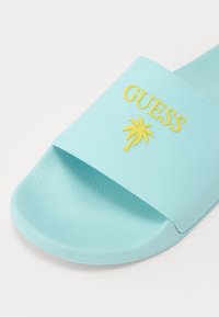 Lys blå gummisandaler med strukturert stropp, med en gul "GUESS" logo og palmegrafikk på den øvre flaten.