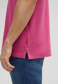 Roze katoenen polo t-shirt met een gestructureerde afwerking, voorzien van zijslits en een rode logo-label aan de zoom, gecombineerd met blauwe spijkerbroeken.