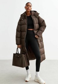 Lång brun puffjacka med huva, öppnad för att avslöja en svart crop top och ribbade leggings. Modellen håller en mörk tote-väska och bär vita sneakers.