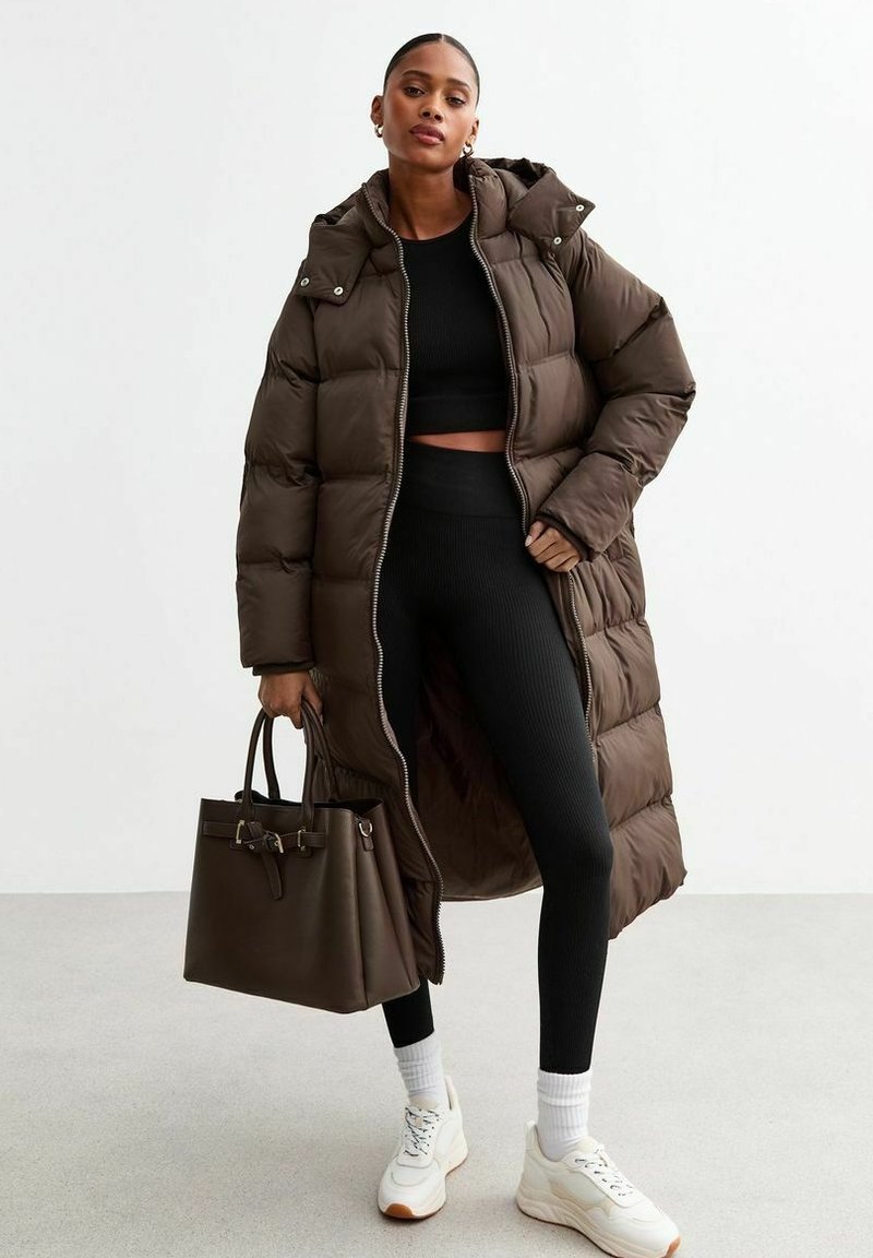 Lång brun puffjacka med huva, öppnad för att avslöja en svart crop top och ribbade leggings. Modellen håller en mörk tote-väska och bär vita sneakers.