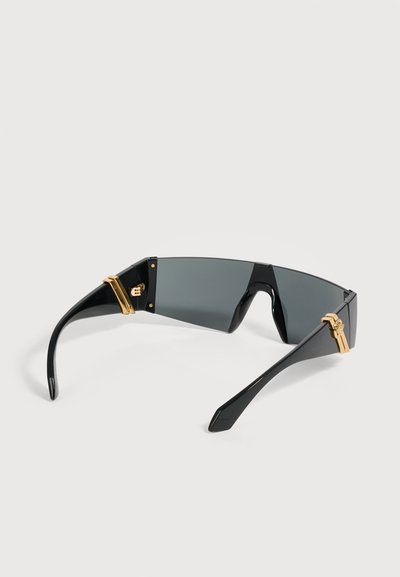Lunettes de soleil enveloppantes noires avec verres rectangulaires foncés et détails dorés aux charnières des branches, présentées sur un fond clair.