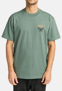 Camiseta de algodón verde con cuello redondo, que presenta un gráfico de una mariposa negra y el texto naranja "RVCA" en el lado izquierdo del pecho. Mangas cortas, corte relajado.