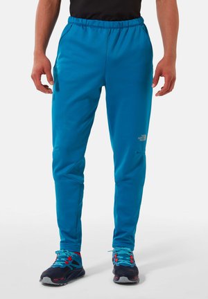Pantalons de sport bleus avec une taille élastique, une texture lisse et des poches latérales, associés à des chaussures noires avec des accents bleu vif et rouge.