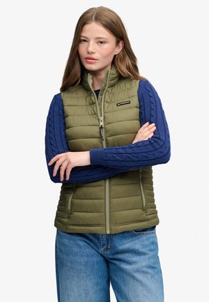 Ung kvinde med langt hår iført en olivengrøn quiltet vest over en blå strikket sweater og blå jeans, med armene krydset.