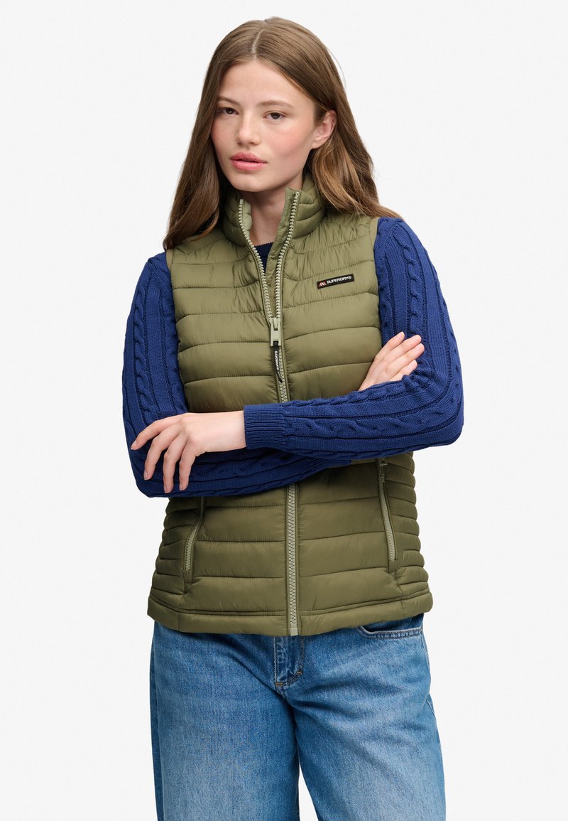 Jeune femme aux longs cheveux portant un gilet matelassé vert olive par-dessus un pull en maille torsadée bleu et un jean bleu, les bras croisés.