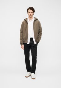 Καφέ φούτερ με κουκούλα πάνω από ένα ανοιχτό ροζ πουκάμισο με κουμπιά, σε συνδυασμό με μαύρο παντελόνι και λευκά αθλητικά παπούτσια με μπεζ σόλες. Απλός σχεδιασμός συνολικά.