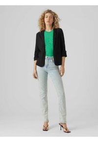 Svart blazer över en grön texturerad topp, ihop med ljusblå raka jeans och svarta remsandaler med hög klack. En enfärgad grå bakgrund.
