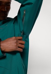 Osoba koja otvara ventilacijski patentni zatvarač ispod pazuha na zelenoj vodootpornoj jakni, s naglaskom na ruku i detalje zatvarača.