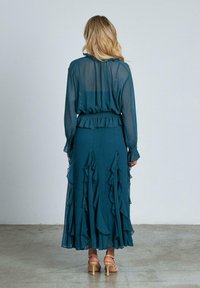 Blusa turchese trasparente con polsini arricciati; gonna lunga coordinata con molte balze; vita aderente. Indossata con sandali con tacco di colore chiaro.