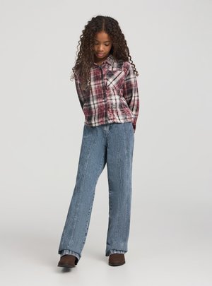 Ragazza giovane con capelli ricci che indossa una camicia a quadri rossa, jeans a gamba larga blu e scarpe marroni, in piedi con la testa leggermente inclinata verso il basso.