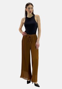 Zwarte mouwloze top gecombineerd met hooggewaardeerde, geplooide roestkleurige wijde broek. Model draagt puntige zwarte hakken.