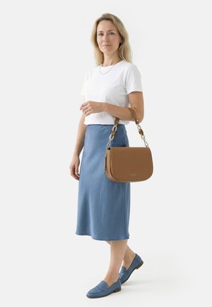 Frau in weißem T-Shirt und blauem Midirock, die eine braune Handtasche mit Kettenhenkel hält und blaue Loafers trägt, vor hellem Hintergrund.