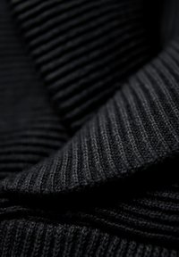 Zwarte ribgebreide stof met een zachte textuur, voorzien van een opvallende kraag en gelaagde plooien, die verschillende diepten van stof laat zien.