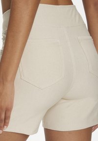 Close-up of tan fabric shorts with back pockets worn by a person with medium skin tone, shown from the waist to mid-thigh.
