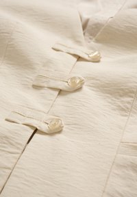 Zbliżenie na beżową tkaninę z trzema przezroczystymi guzikami przyszytymi do pasujących pętelek z tkaniny.