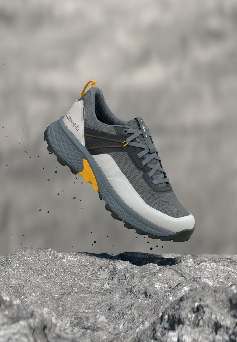 Γκρι και λευκό παπούτσι πέδης Columbia με κίτρινα στοιχεία, αιωρούμενο πάνω από πετρώδες έδαφος, με σκόνη και μικρές πέτρες να πέφτουν από κάτω του.