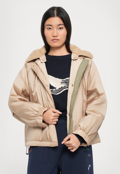 Veste matelassée beige avec un col en fausse fourrure marron amovible, des détails intérieurs verts et des poignets élastiques, portée sur un pull bleu foncé.