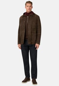 Man som bär en brun mockajacka, mörkbrun zip-up-tröja, mörkblå jeans och bruna skor, som står mot en enfärgad bakgrund.