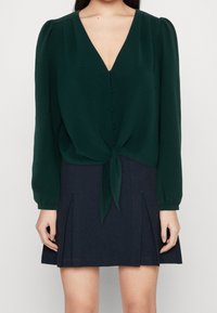 Blusa verde scuro con scollo a V, maniche lunghe e dettagli con bottoni, legata in vita, abbinata a una gonna plissettata blu navy.