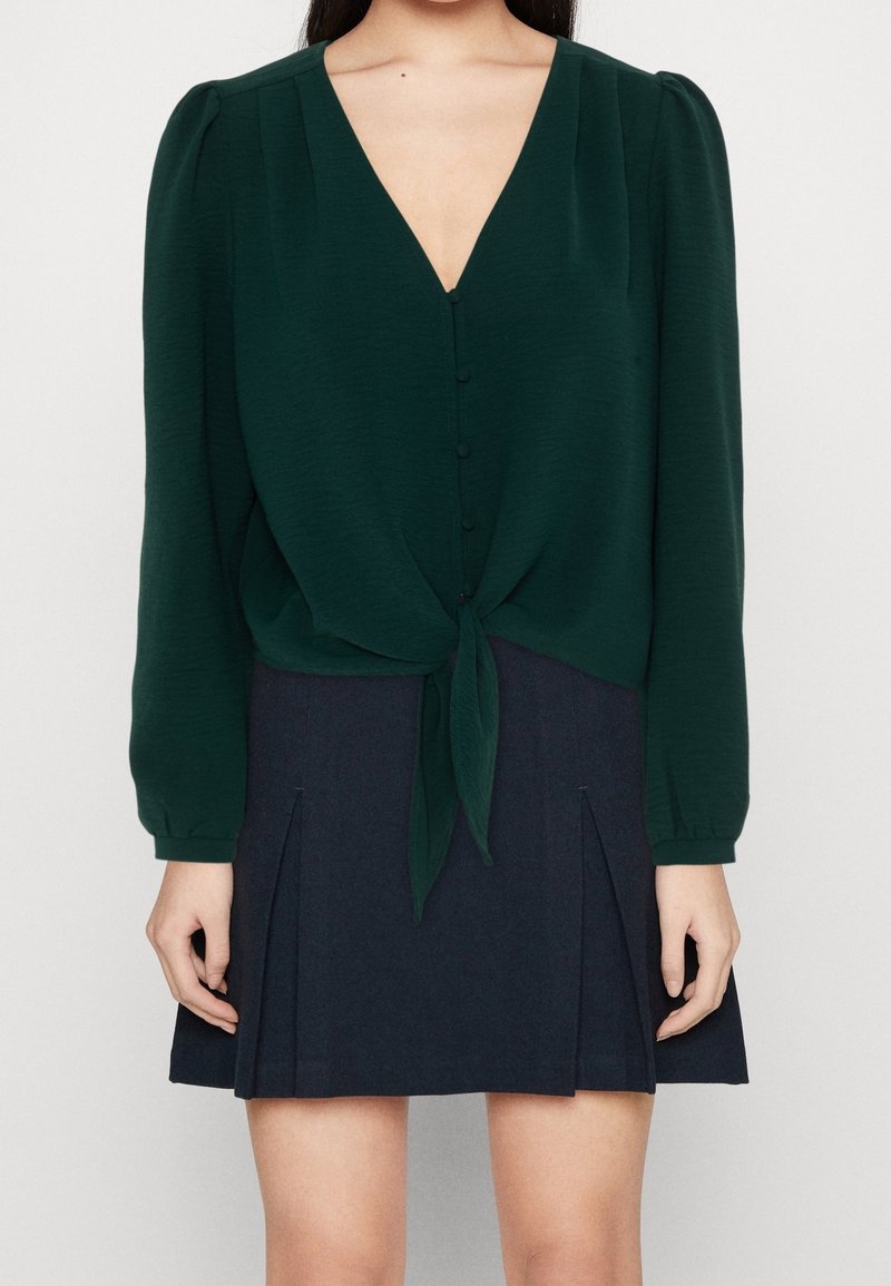 Blusa verde scuro con scollo a V, maniche lunghe e dettagli con bottoni, legata in vita, abbinata a una gonna plissettata blu navy.