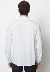 Camicia bianca a maniche lunghe con colletto a punta, polsini con bottoni e texture liscia; indossata con jeans scuri, presenta un orlo dritto.