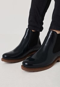 Mustad musta nahast pahkluusaapad elastsete külgpaneelidega ja pruunist kummist tallaga. Sujuv tekstuur, ümmargune nina kujundus ja minimaalne detaile.