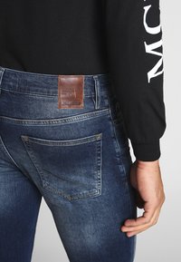 Jeans de gama oscura con un acabado texturizado. Presenta un parche de cuero marrón en la cintura trasera y bolsillos traseros estándar. Camisa negra de manga larga.