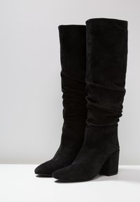 Svarta mockakängor i knähöjd med en avslappnad design, spetsig tå och kraftig klack. Slät yta utan synliga mönster eller detaljer.