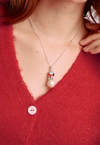 Silberne Halskette mit einem Anhänger im Charakterdesign, roten Akzenten und einem Perlenanhänger, getragen mit einer roten strukturierten Strickjacke.