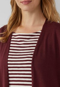 Bordeauxfarbener Cardigan mit offenem Vorderteil, getragen über einem gestreiften Shirt mit roten und weißen horizontalen Streifen, aus weichem Stoff und mit lockerer Passform.